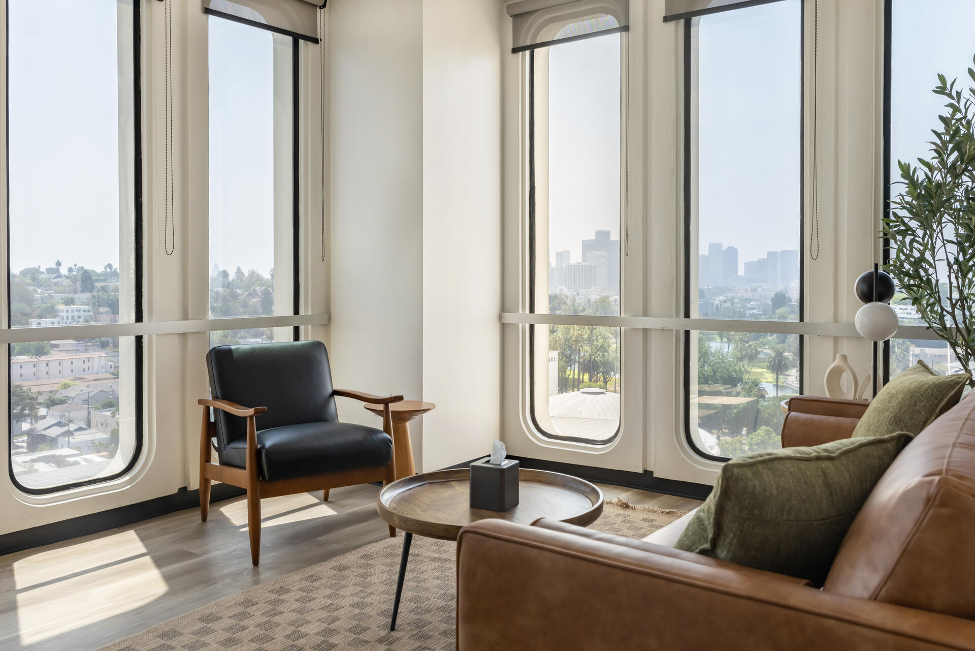 Therapy office in Echo Park — mid-century leather chair, warm sofa, and floor-to-ceiling windows overlooking Echo Park Lake and the downtown Los Angeles skyline